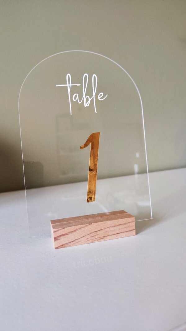 Numéros de table pour mariage, en acrylique transparent avec calligraphie dorée placés sur une table décorée.