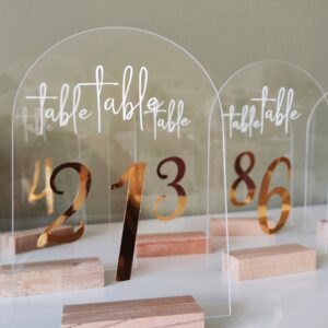 Numéros de table pour mariage, en acrylique transparent avec calligraphie dorée placés sur une table décorée.