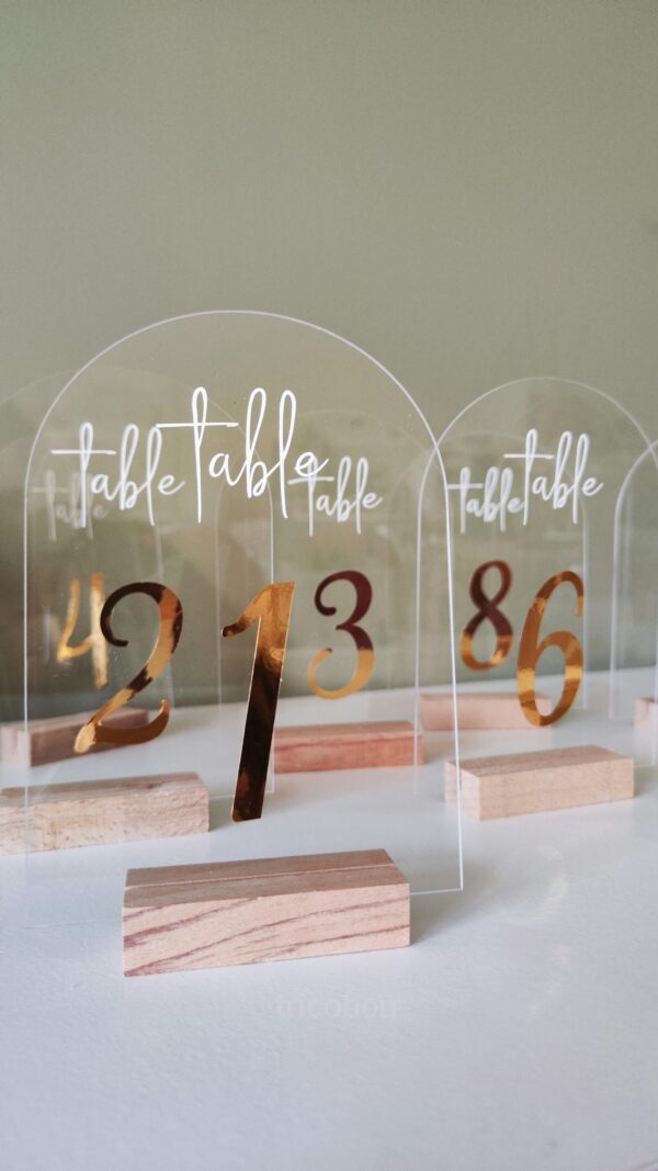 Numéros de table pour mariage, en acrylique transparent avec calligraphie dorée placés sur une table décorée.